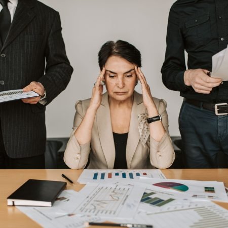 Dott. Cristiano Pedersini - Donna preoccupata alla scrivania circondata da colleghi, simbolo di sovraccarico, pressione psicologica e senso di invisibilità, consulenza per il burnout